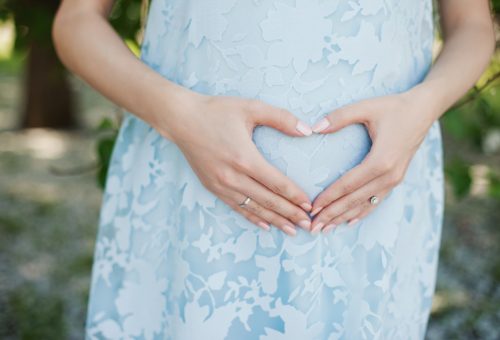 Pregnant woman in light blue dress holding her pregnant belly and love sign - maternity concept